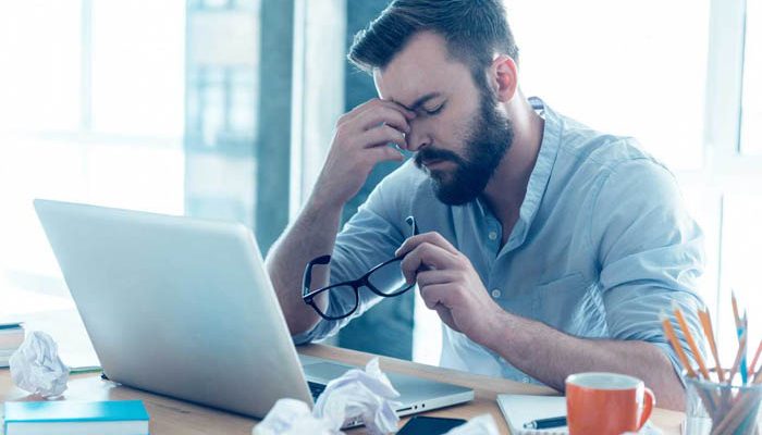 Frustrated young beard man massaging his nose and keeping eyes closed while sitting at his working place in office